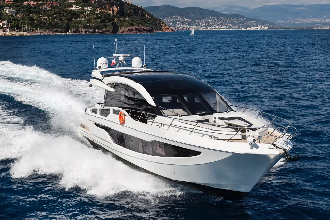 A sleek motor yacht gliding through clear blue water with scenic hills and a distant shoreline in the background.