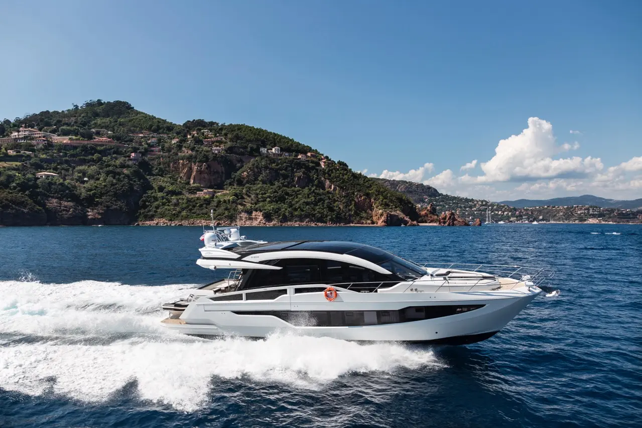 A modern motor yacht cuts through blue waters with a scenic hillside and coastal homes in the background under a clear sky.