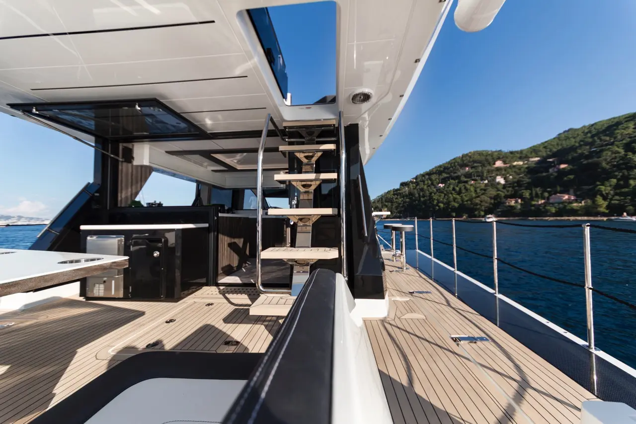 Luxury yacht deck with sleek design, stairs leading up, outdoor seating, and stunning water views of a lush green landscape.