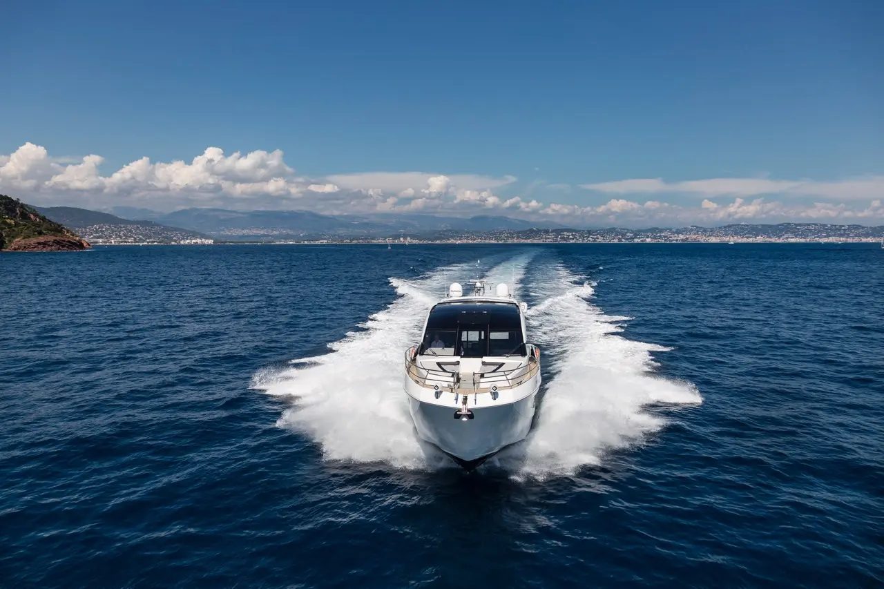 A sleek motorboat navigates smooth blue waters, leaving a white wake against a backdrop of hills and distant coastline.