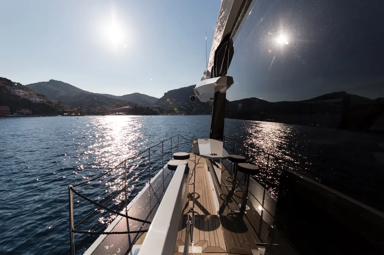 View from a yacht’s deck showing sunlight on water, mountain backdrop, and stylish stools on a wooden surface.
