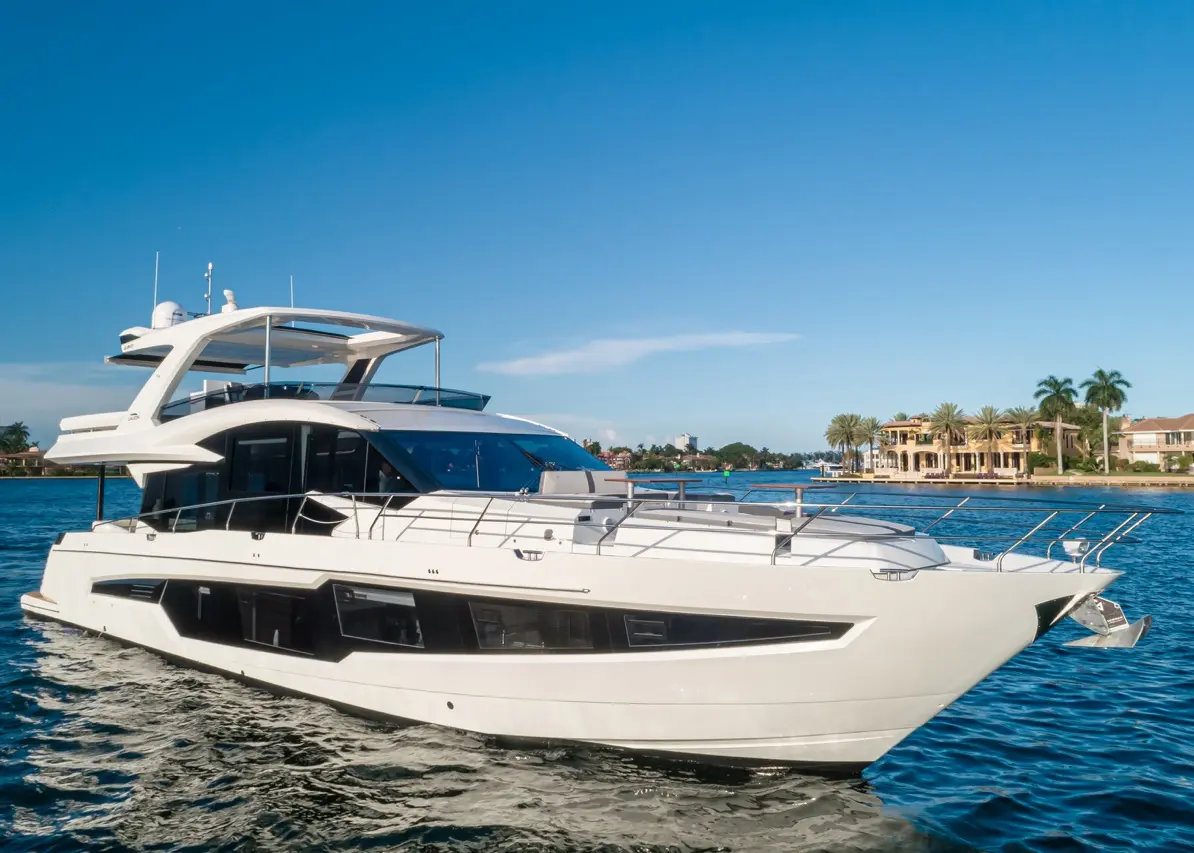 A sleek white yacht glides through calm waters, with luxurious homes and palm trees visible in the background under a clear blue sky.