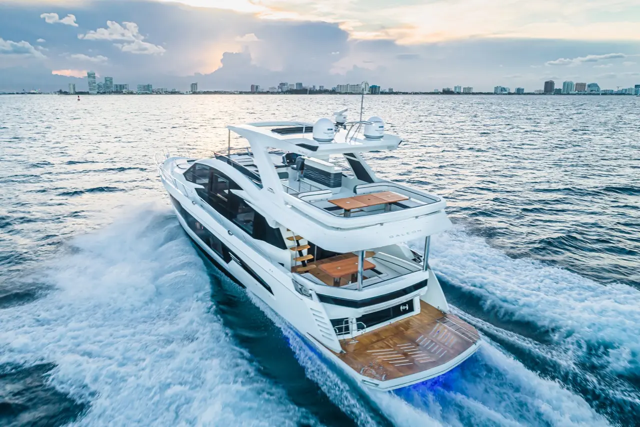 Luxury yacht cruising on calm waters, leaving a wake, with a city skyline visible under a sunset sky.
