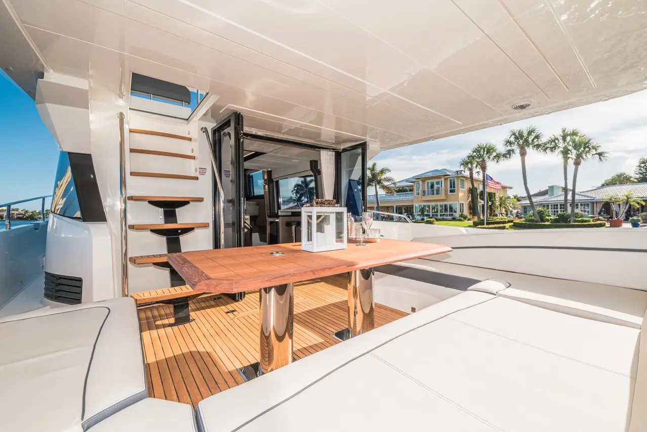 Stylish yacht interior featuring a wooden table, comfortable seating, and stairs leading to the upper deck, with palm trees visible outside.