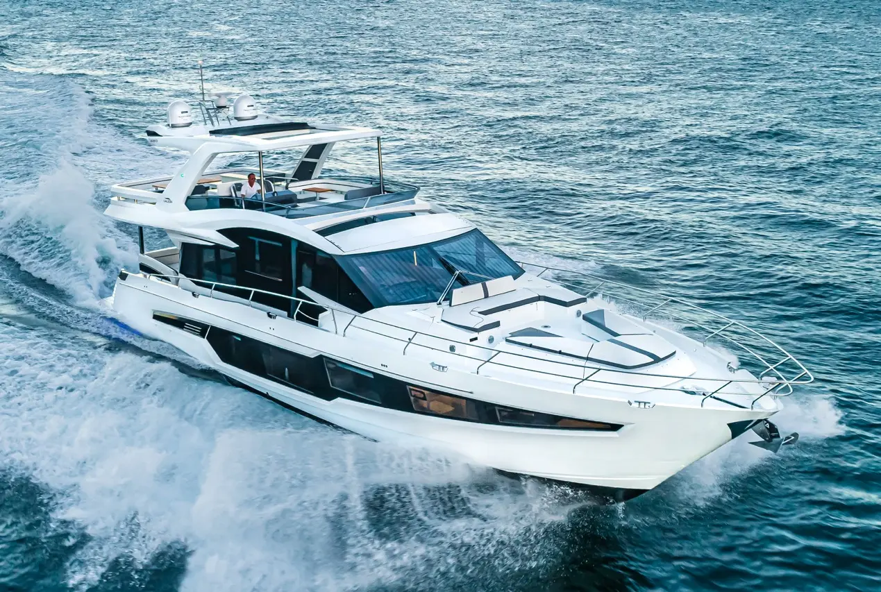 A sleek white yacht cruising through blue waters, leaving a wake behind; a person is visible on the upper deck.