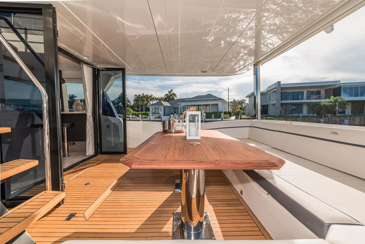 Luxury yacht deck with a wooden dining table, featuring a clear skyline view and plush seating. Coastal homes are visible.