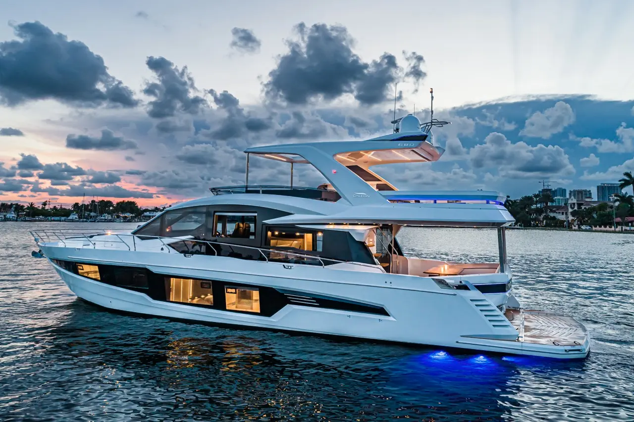 A sleek white yacht with blue LED lights moored on calm waters during sunset, surrounded by palm trees and cloudy sky.