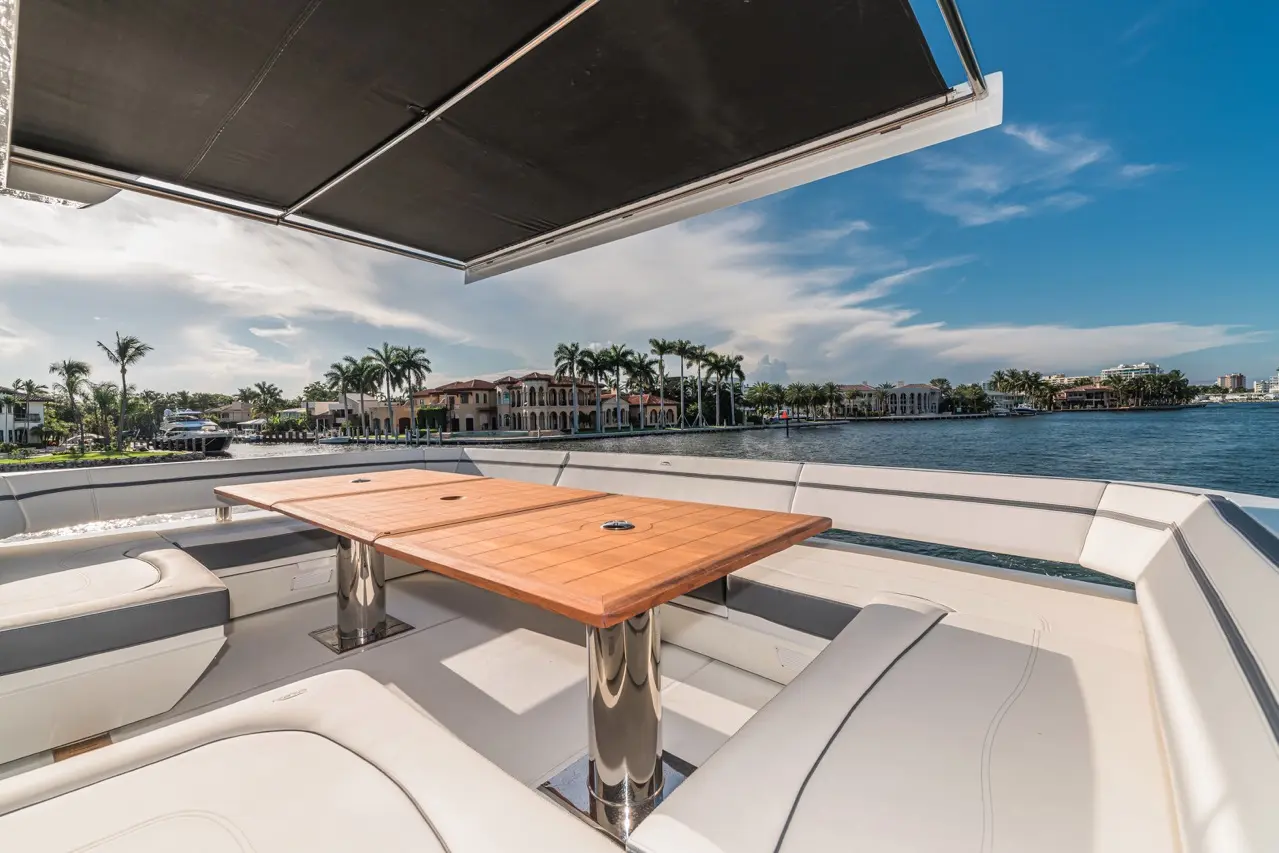 Luxury boat deck with a wooden table, plush seating, and views of waterfront homes and palm trees under a blue sky.