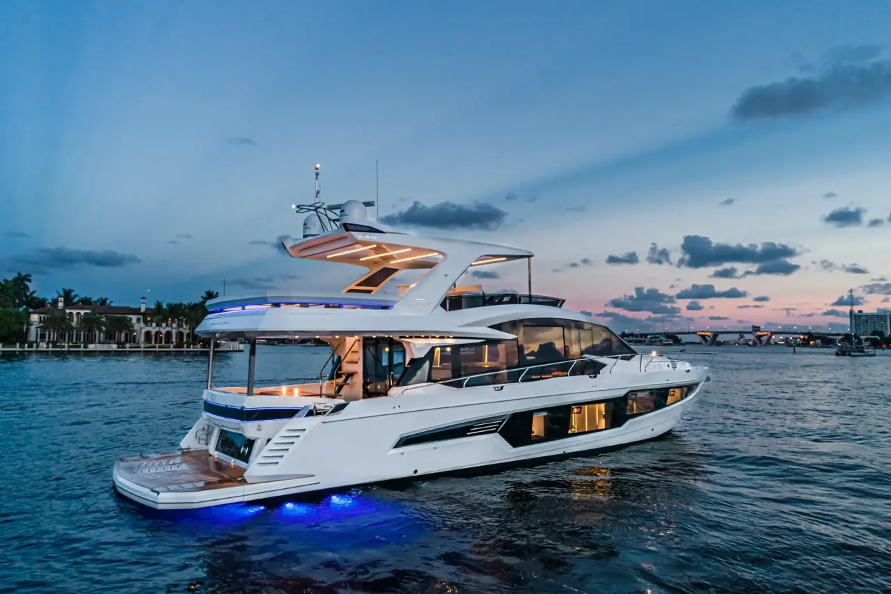 Luxurious yacht illuminated at dusk, gliding over calm waters with palm trees and upscale homes in the background.