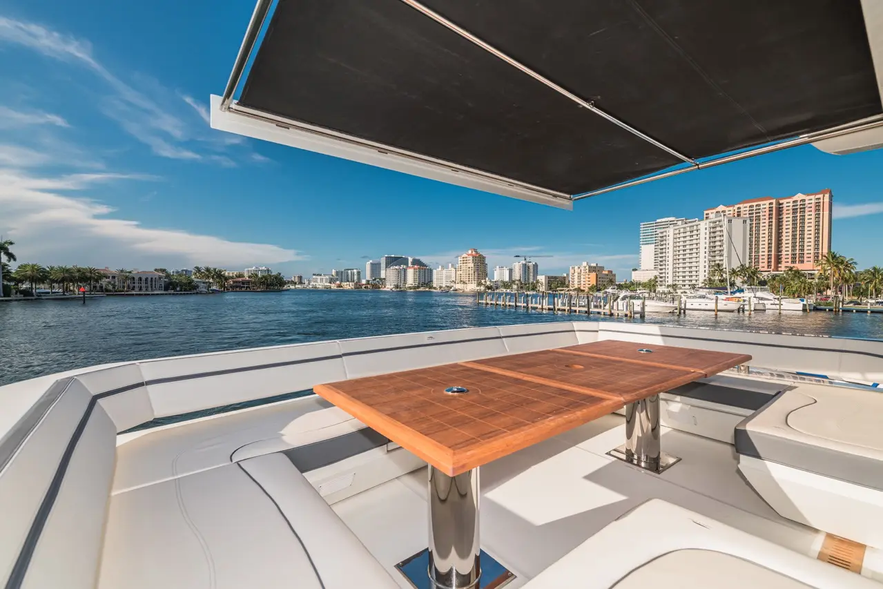 Luxury yacht deck view featuring a polished wooden table, grey seating, and a waterfront skyline under a clear blue sky.
