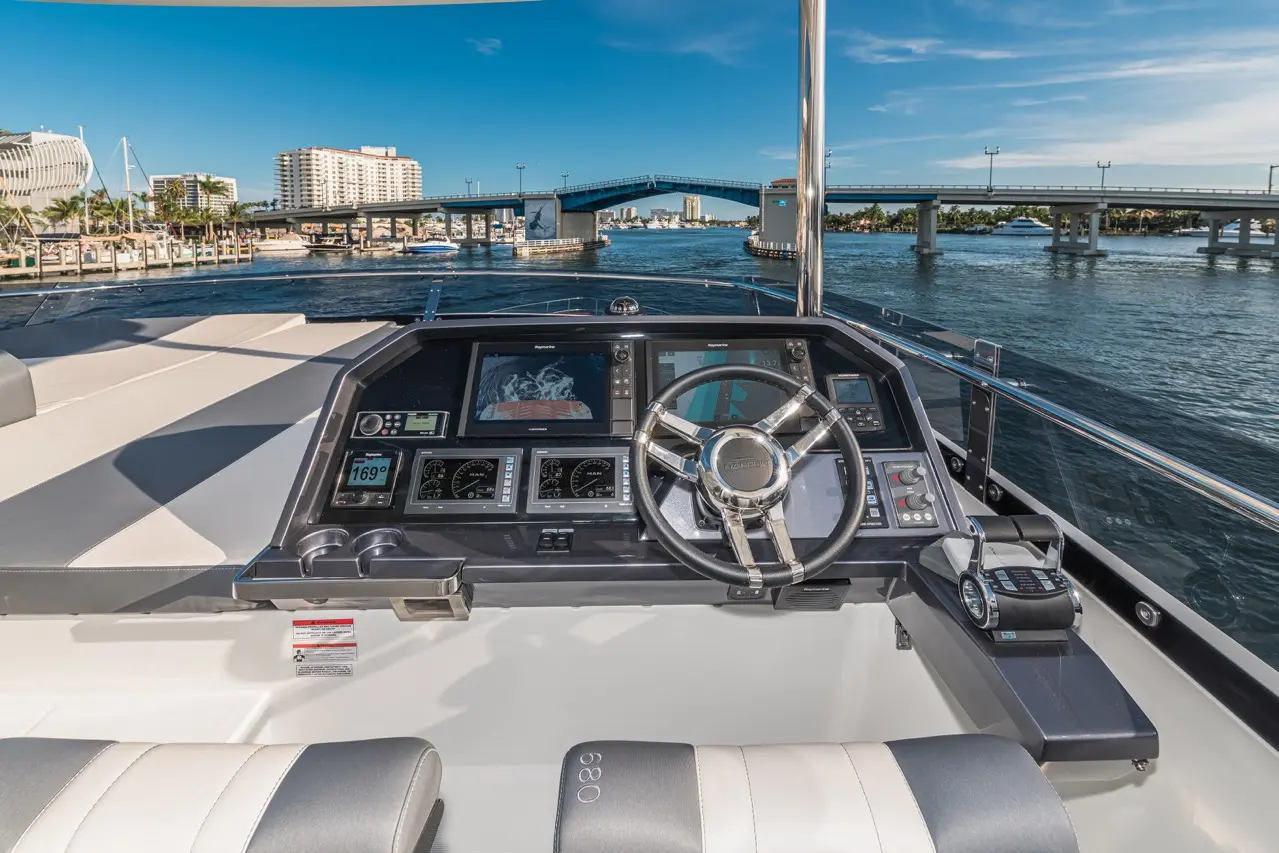 Boat cockpit view featuring a steering wheel, navigational screens, and a scenic waterfront bridge in the background.