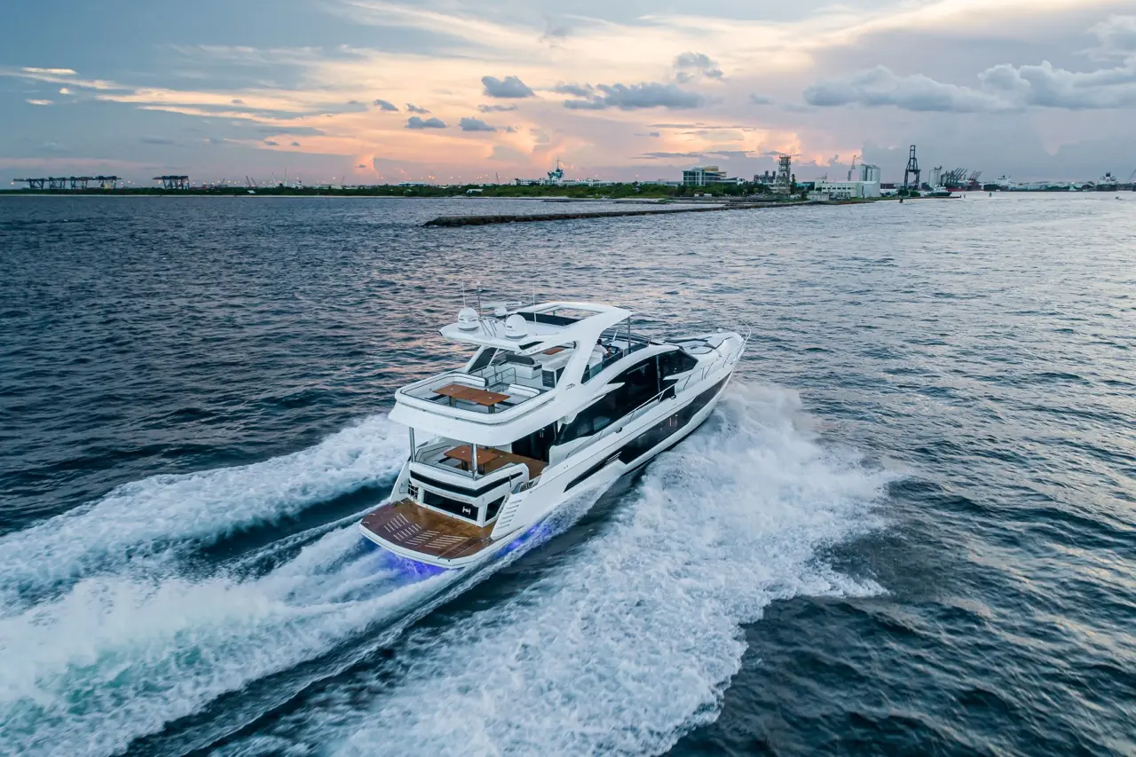 A luxury yacht cruises through calm waters at sunset, creating waves, with a coastal silhouette in the background.