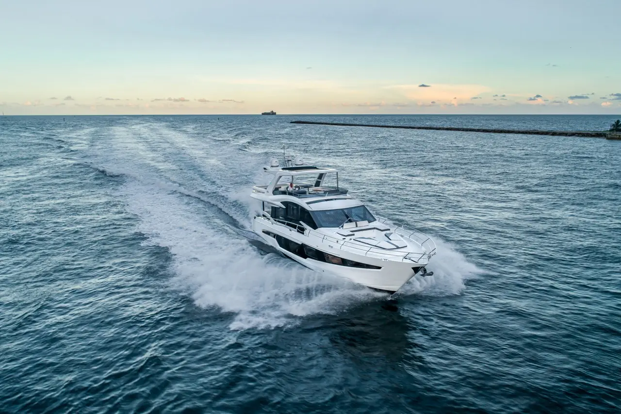 A luxury motor yacht speeds through calm waters, leaving a wake behind against a scenic sky at dusk.