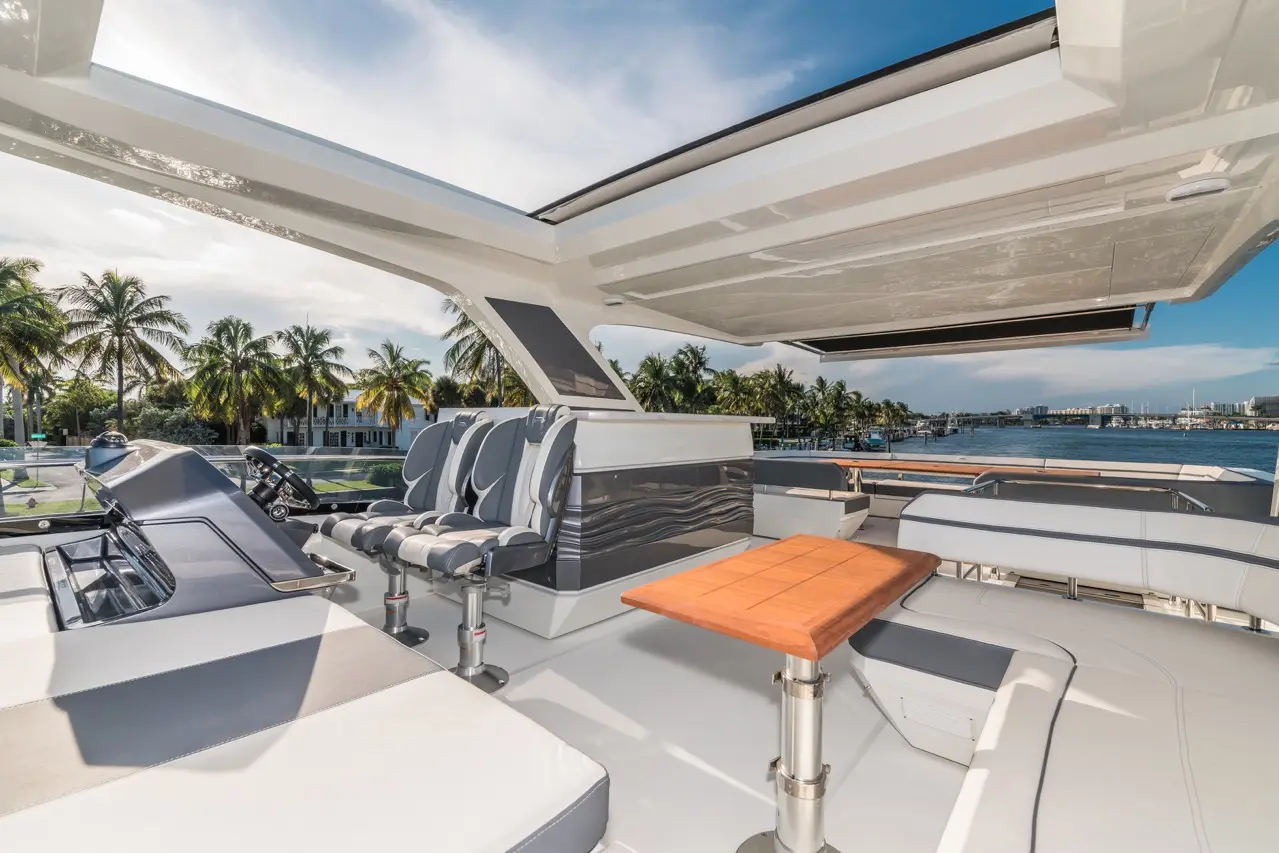 Stylish yacht interior with grey and white seats, wooden table, and palm trees by a waterfront under a clear blue sky.