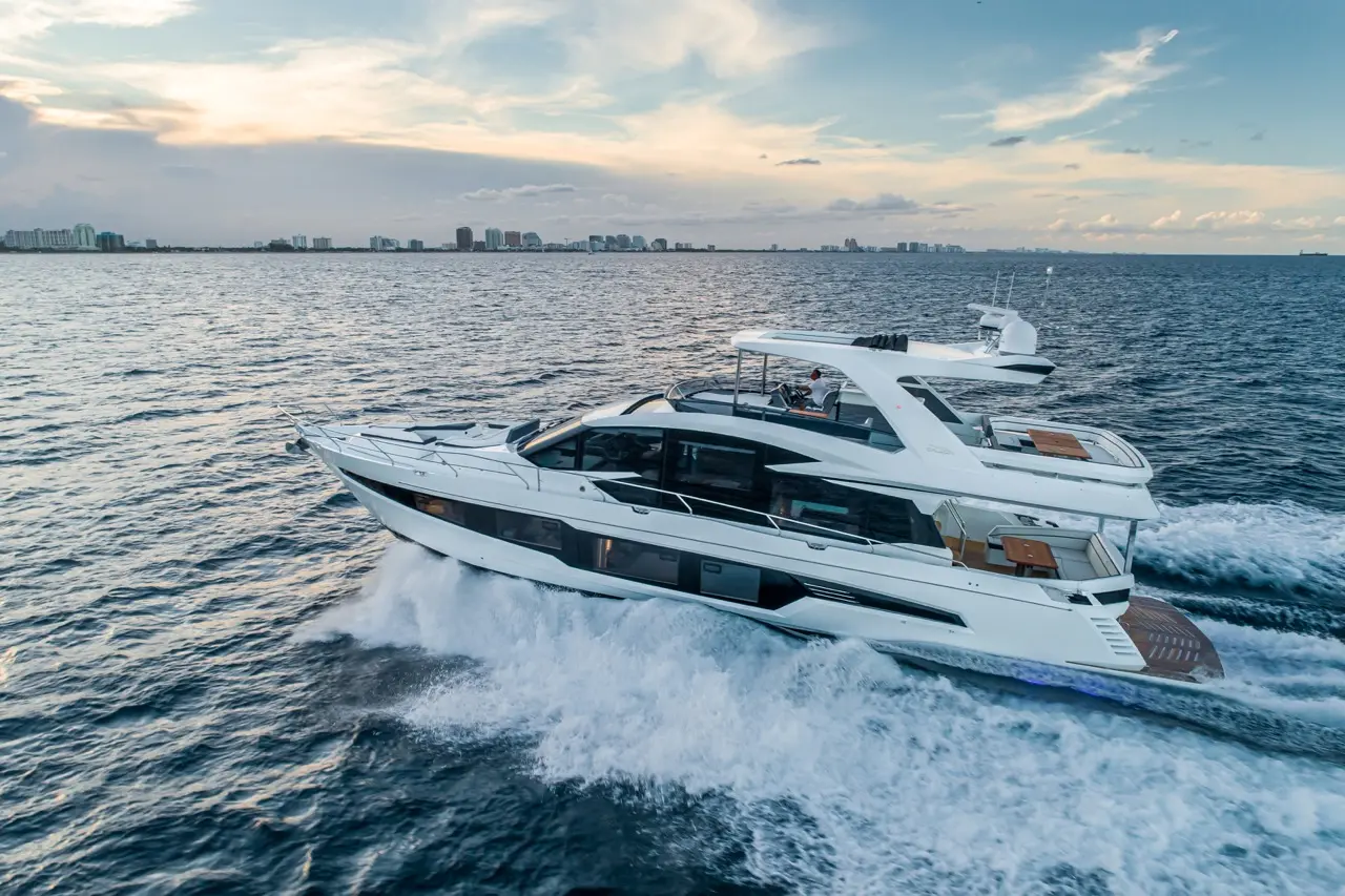 A luxury motor yacht cuts through ocean waves under a colourful sky, with urban skyline visible in the background.