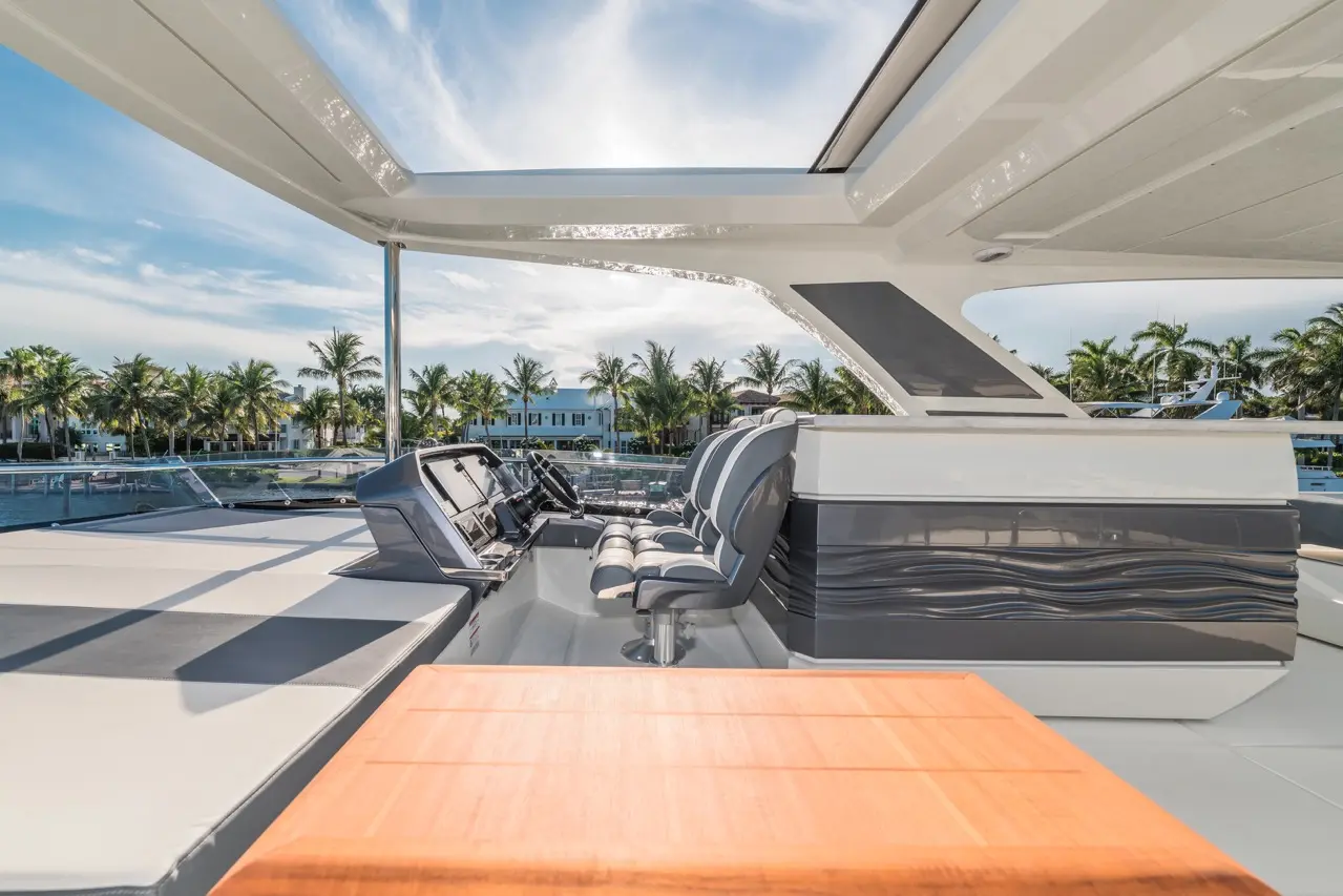 Luxury yacht interior features a modern helm with grey seating, a wooden table, and views of palm trees and water.