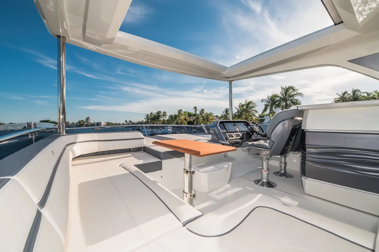 Light-filled yacht cockpit featuring a wooden table, modern controls, and plush seating, with palm trees and water in the background.