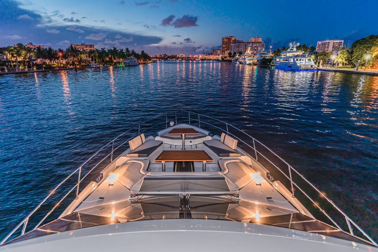 View from the bow of a luxury yacht at dusk, overlooking a calm waterway with city lights reflecting on the water.