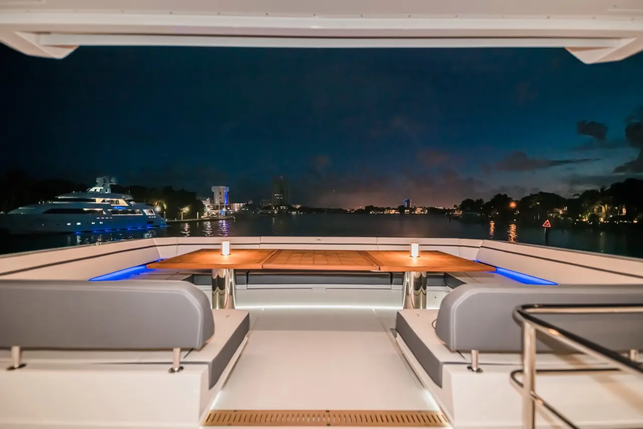 Luxury yacht deck at night with a wooden table, grey seating, and views of city lights reflecting on water.