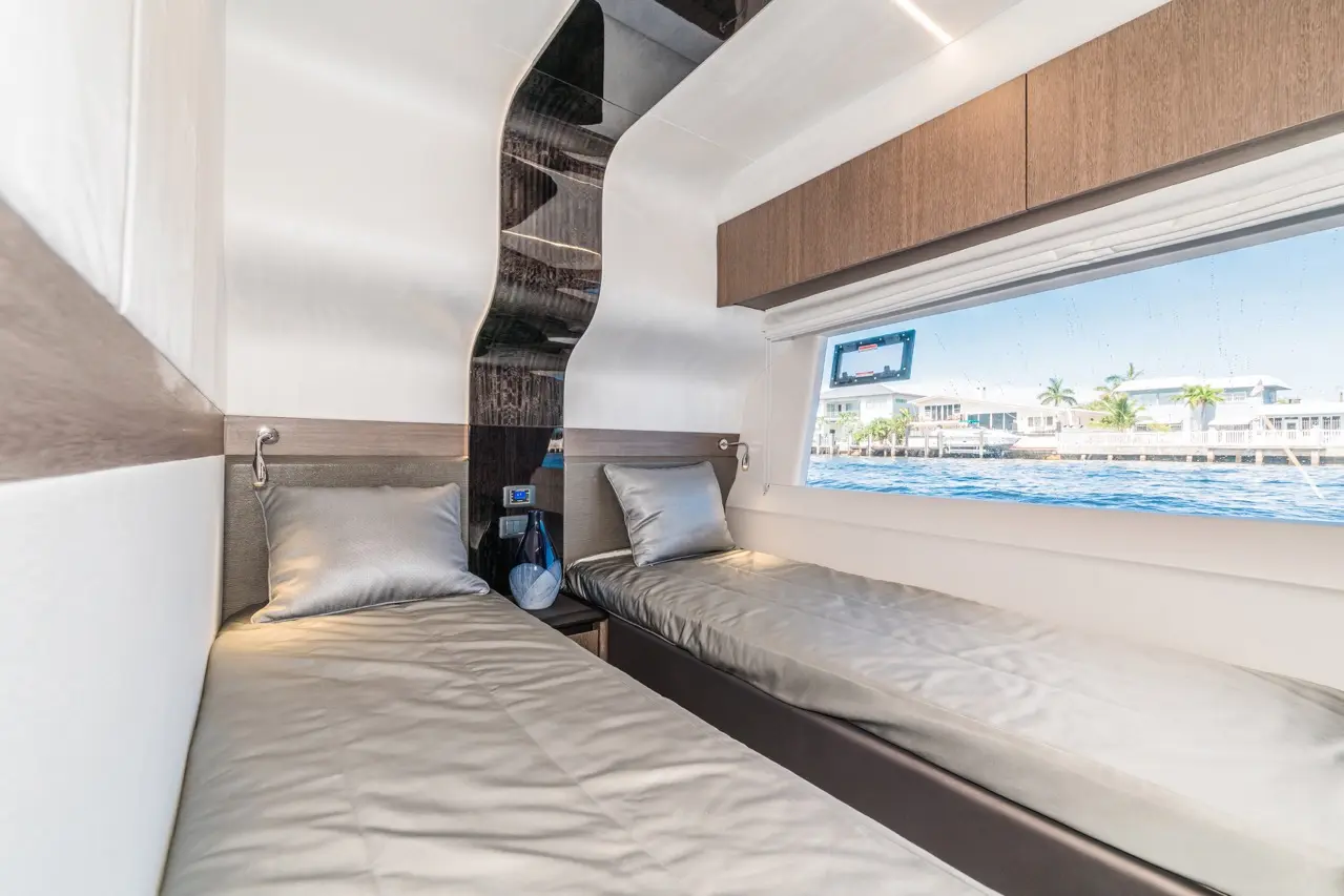 A modern boat cabin featuring two single beds with grey bedding, a window view of waterfront homes, and sleek wooden accents.