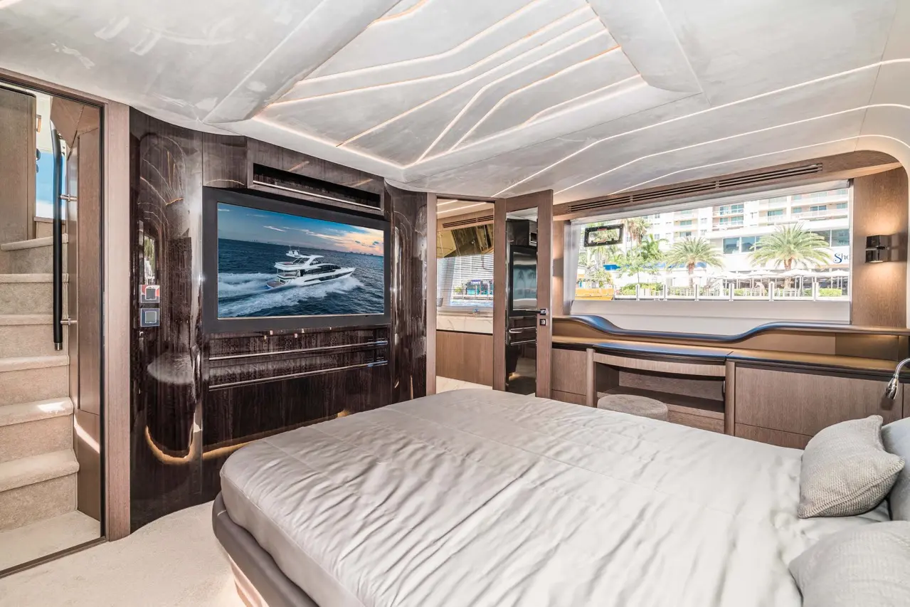 Luxurious yacht bedroom featuring a large TV displaying a yacht, elegant decor, stairs, and windows with a coastal view.