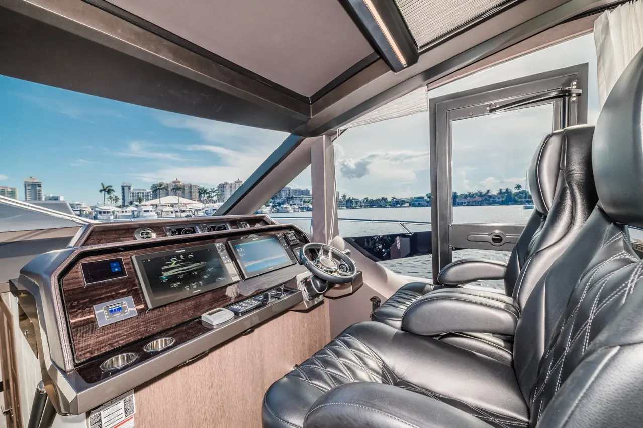 Modern yacht cockpit with dual screens, steering wheel, and comfortable leather seating; scenic marina view through large windows.