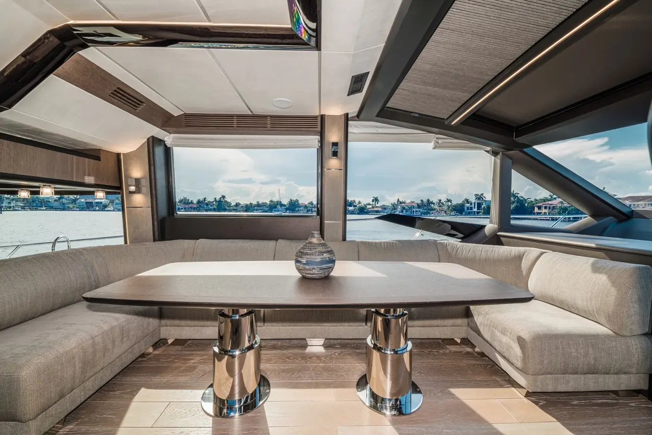 Luxurious yacht interior featuring a grey seating area, a wooden table, and scenic views of the waterfront through large windows.