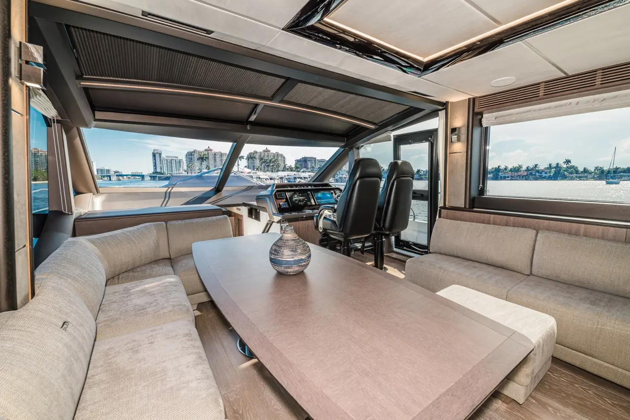 Modern yacht interior featuring a plush seating area, a central table, and a helm with twin black chairs, with waterfront views.
