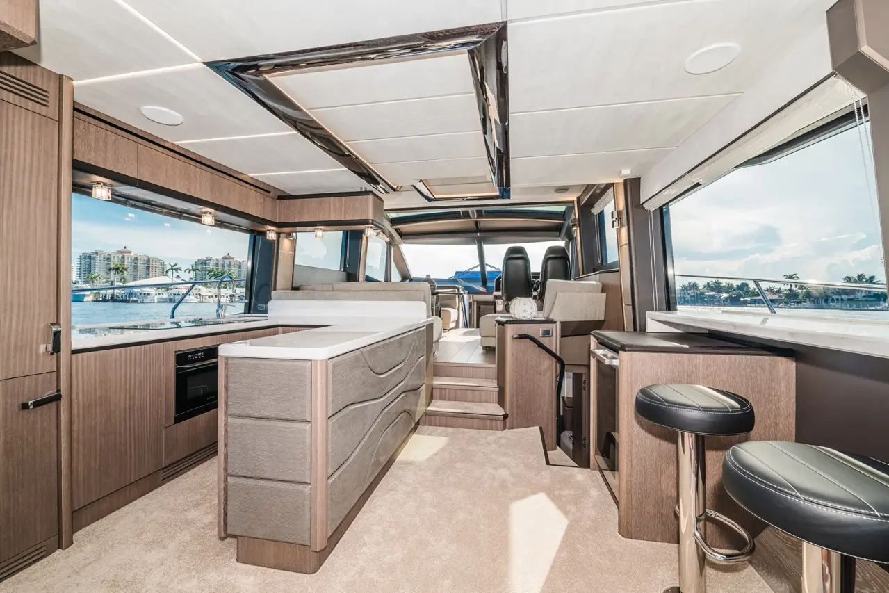 Spacious yacht interior with modern furnishings, panoramic windows, steering area, and a kitchen island, showcasing a waterfront view.