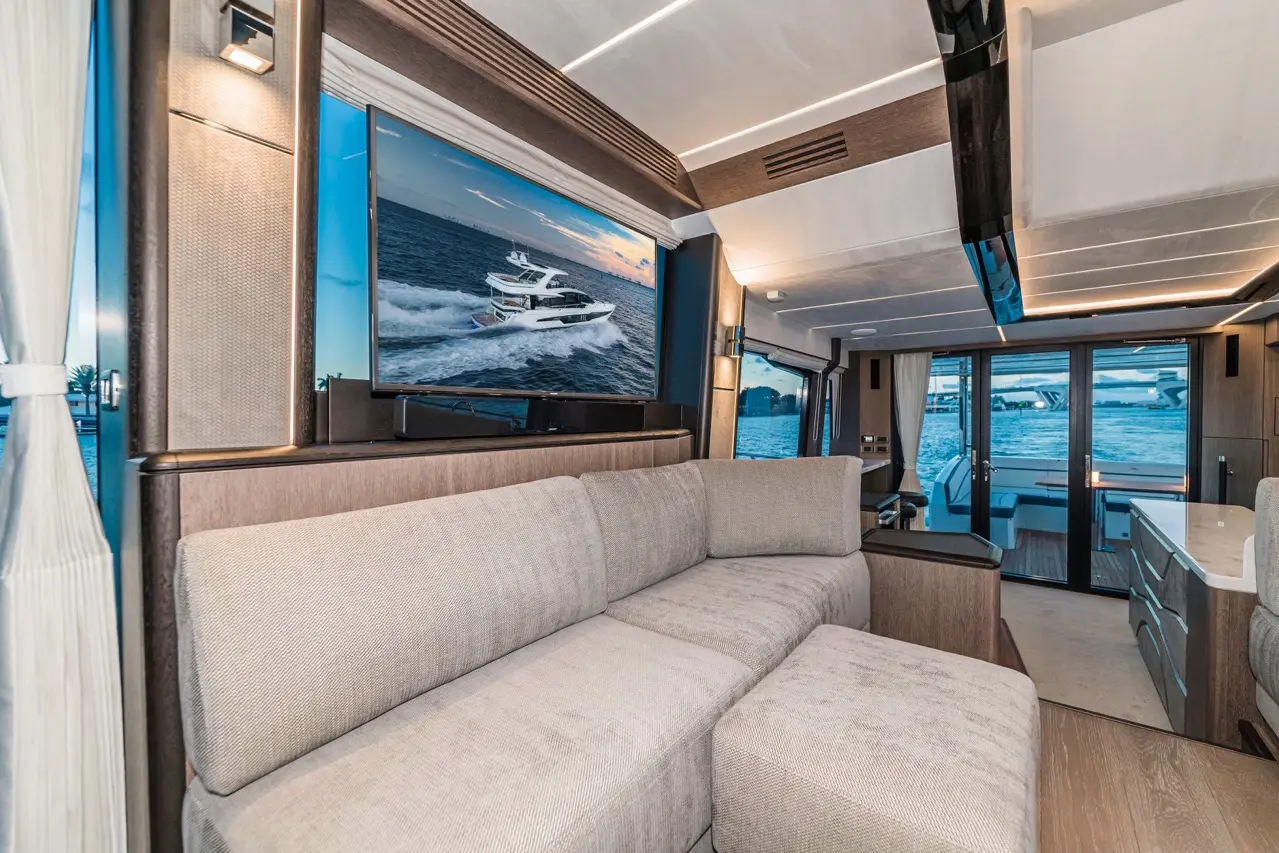 Modern yacht interior featuring a plush beige sofa, a large TV displaying a boat on water, and large windows with sea views.