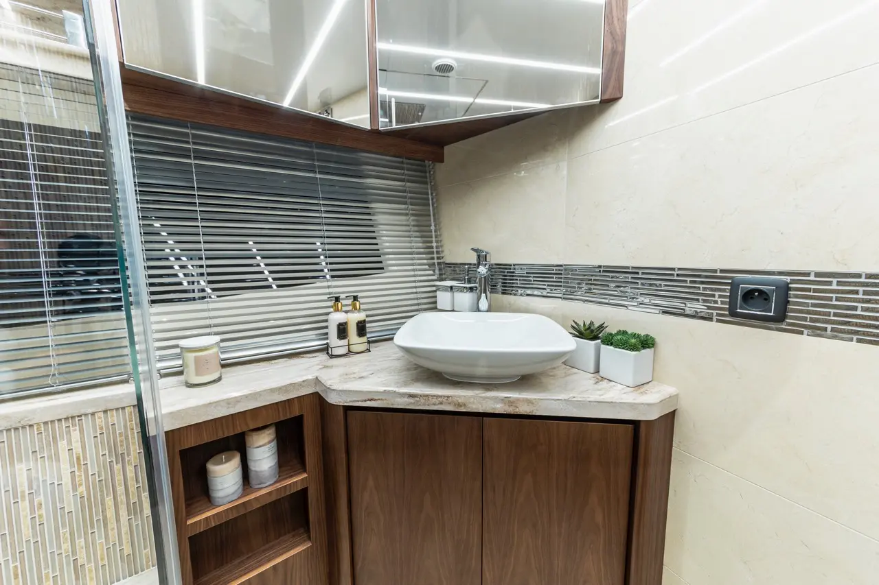 Modern bathroom with a white basin, wooden cabinetry, mirror, decorative plants, and candles on a marble countertop.
