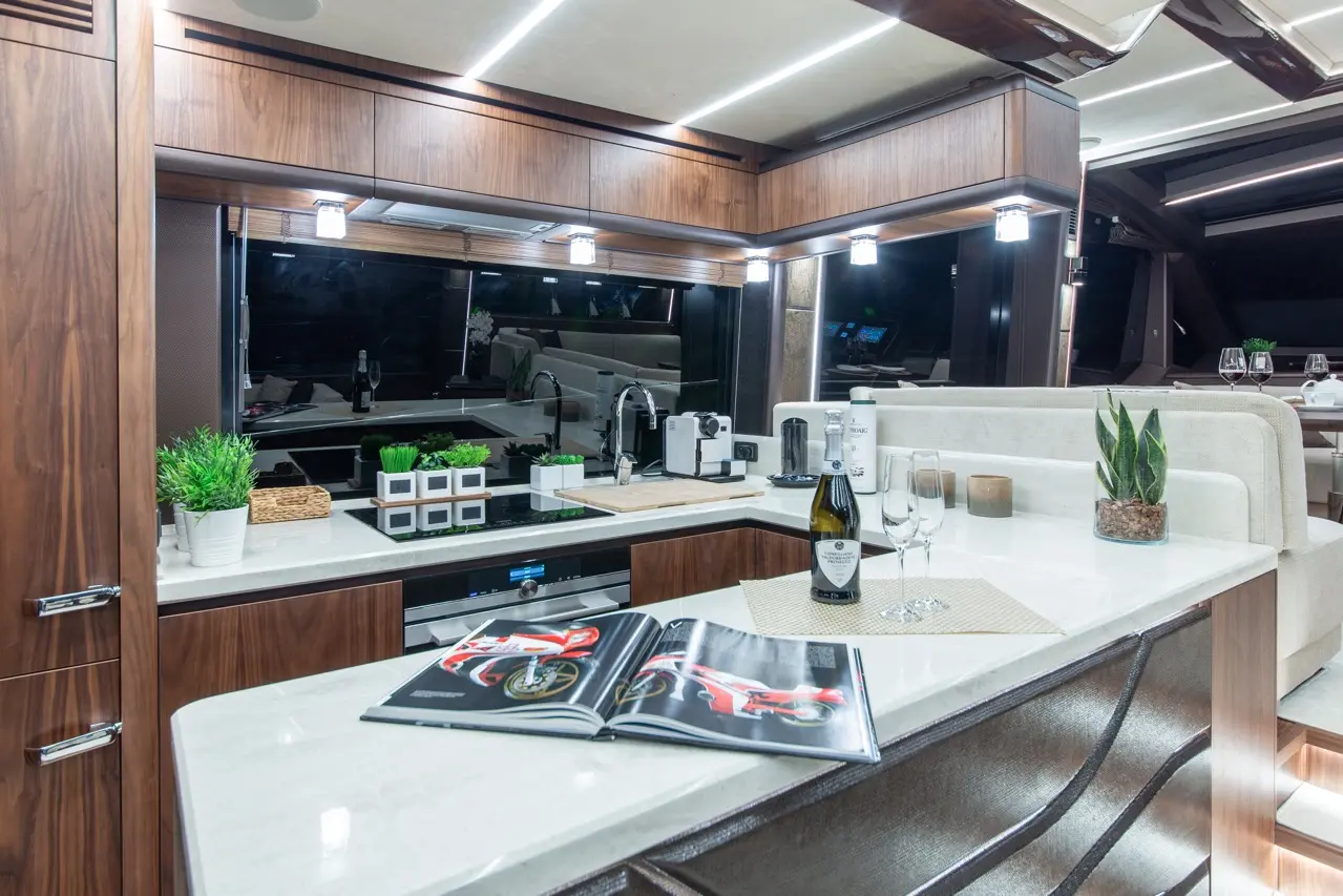 Modern yacht kitchen featuring wooden cabinets, wine, glasses, plants, and an open magazine on a white countertop.