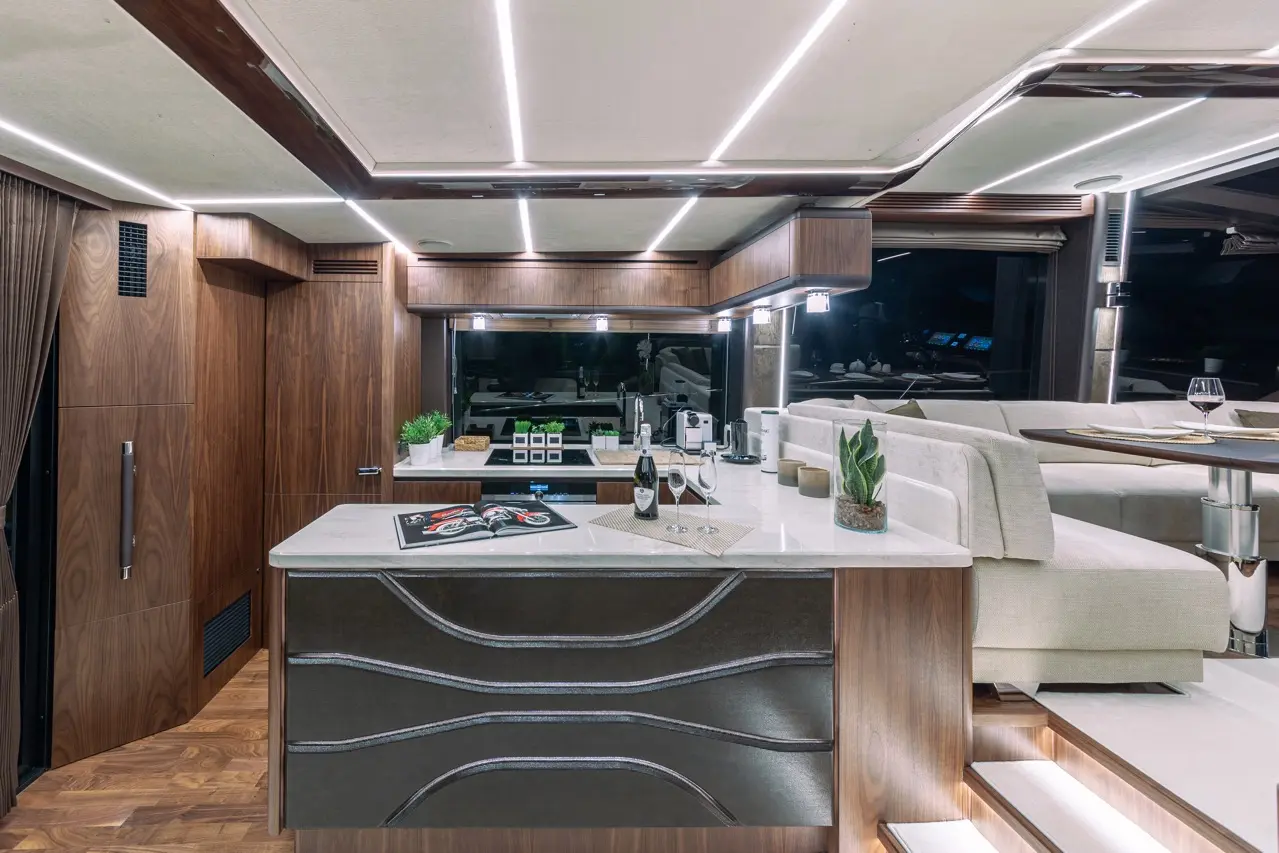 Luxury yacht kitchen with sleek wooden cabinetry, a marble island, wine glasses, and modern appliances, illuminated by LED lights.