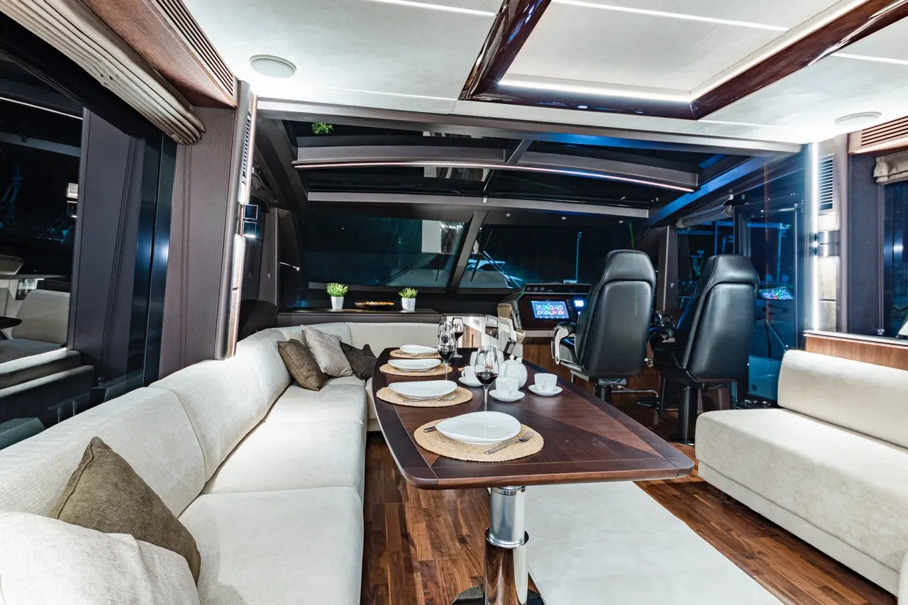 Luxurious yacht interior with a plush sofa, dining table set for dinner, and a modern helm station visible through large windows.