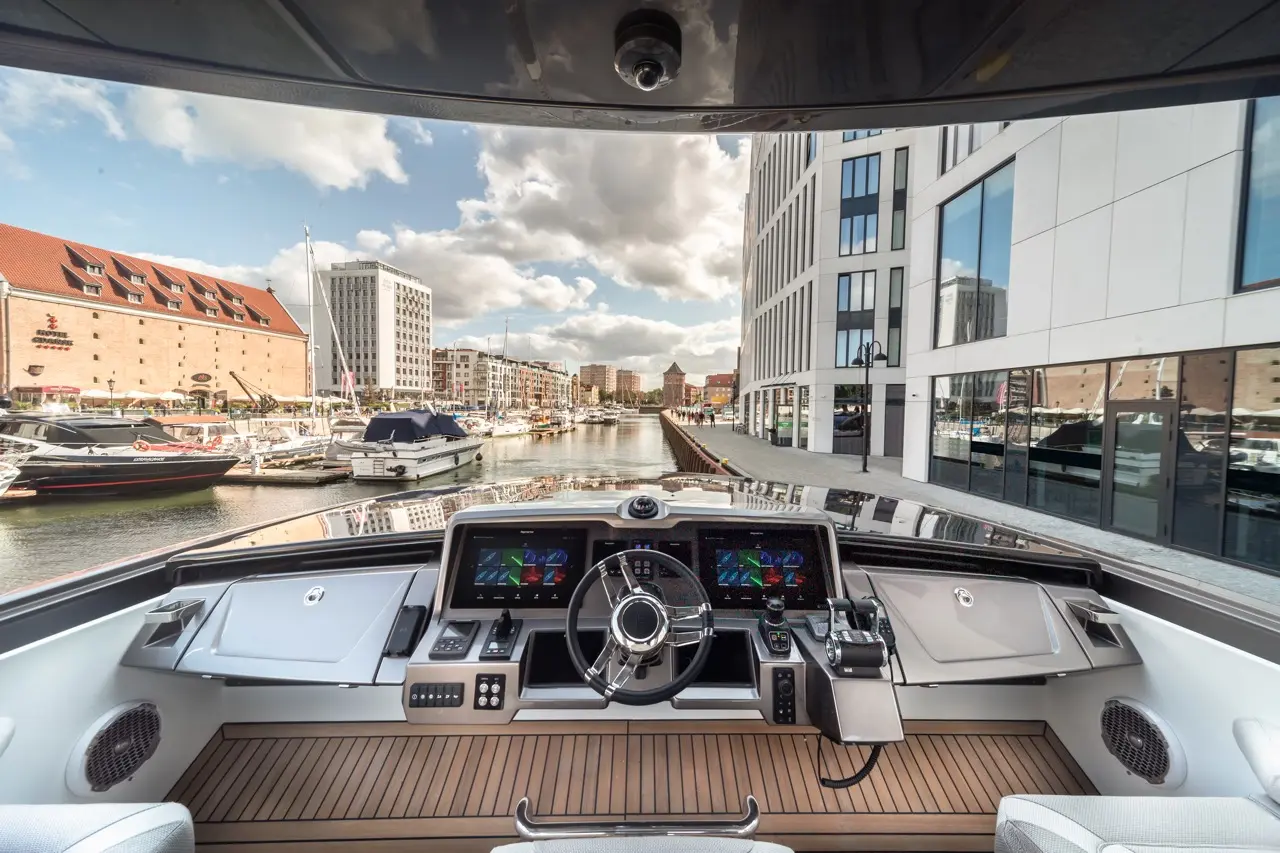 View from a luxury boat's helm, showing modern controls, waterfront buildings, and boats moored along a canal.