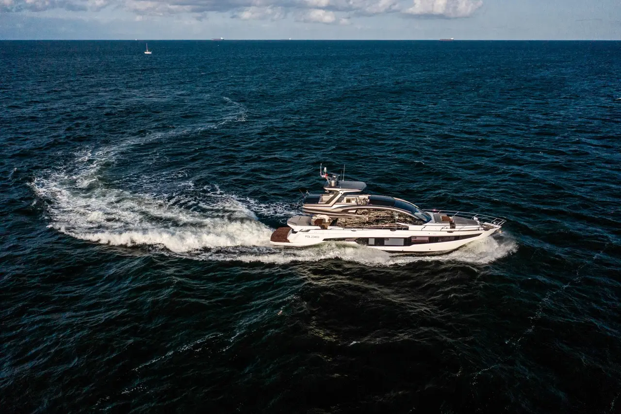 A luxury yacht cruising over deep blue waters, leaving a white wake behind, with distant boats visible on the horizon.