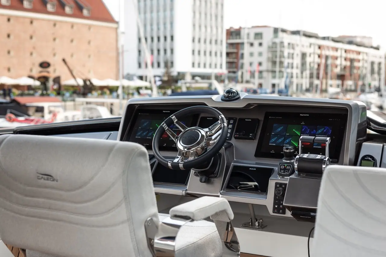 Modern yacht cockpit featuring a steering wheel, digital displays, and navigation controls, with a marina view in the background.