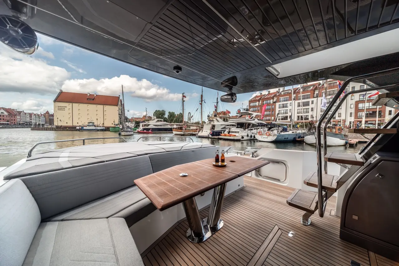 Luxury boat interior featuring a wooden dining table, two drinks, and a view of the marina with moored yachts and buildings.
