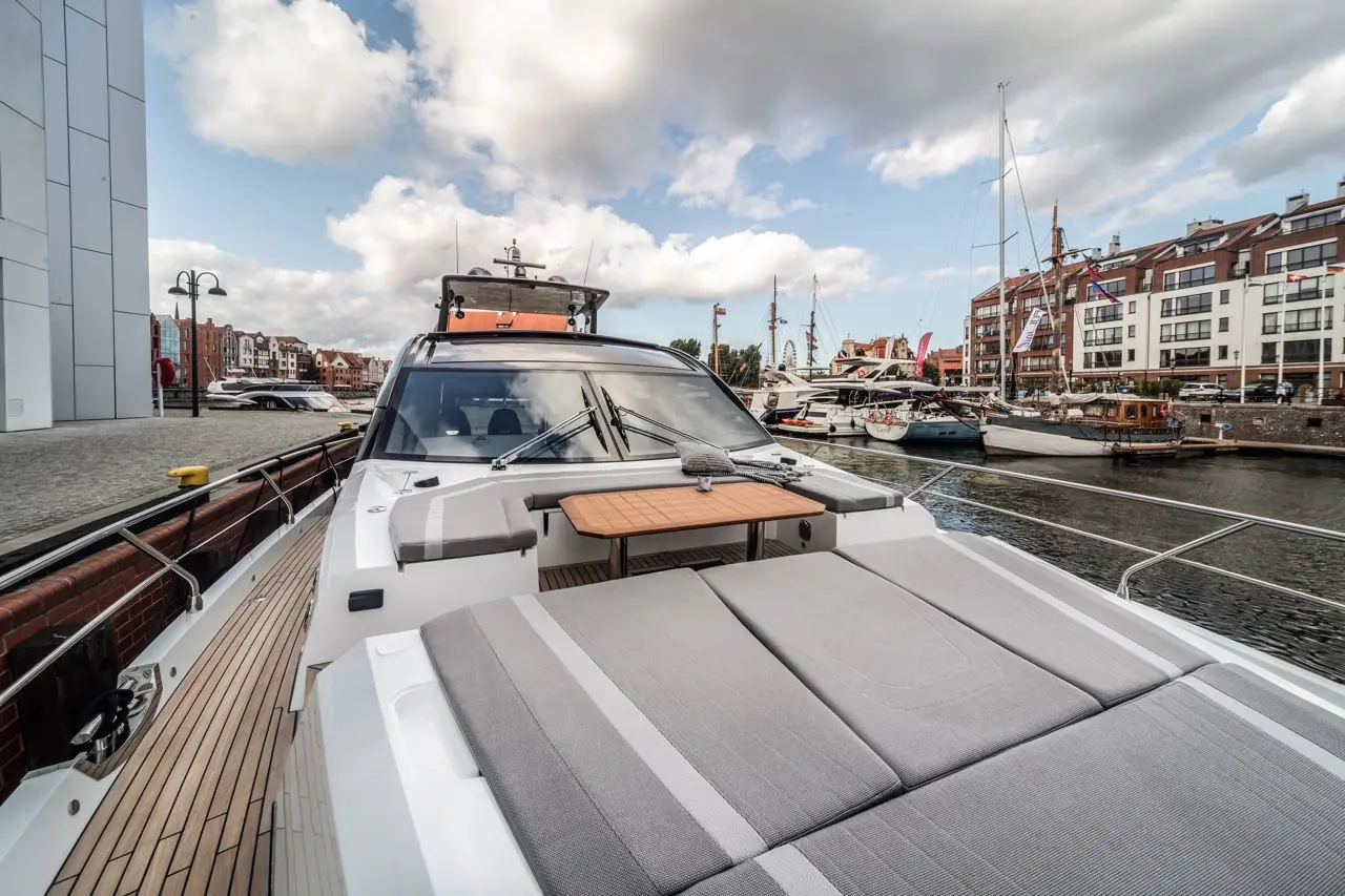 A view from the bow of a modern yacht, showcasing grey seating, a wooden table, and a marina with various boats.