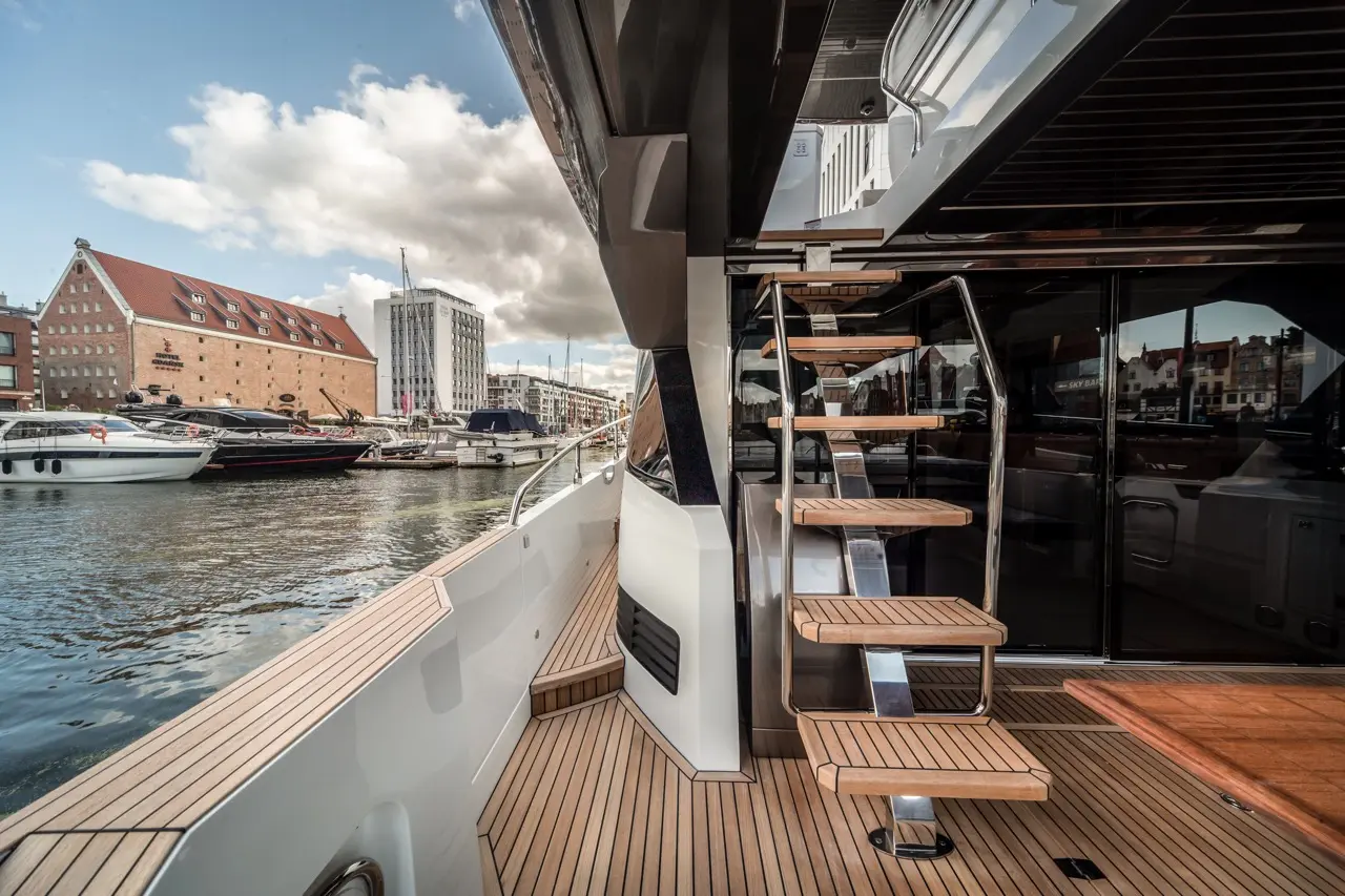 View of a luxurious yacht's deck with wooden stairs, overlooking a marina filled with boats and striking architecture.