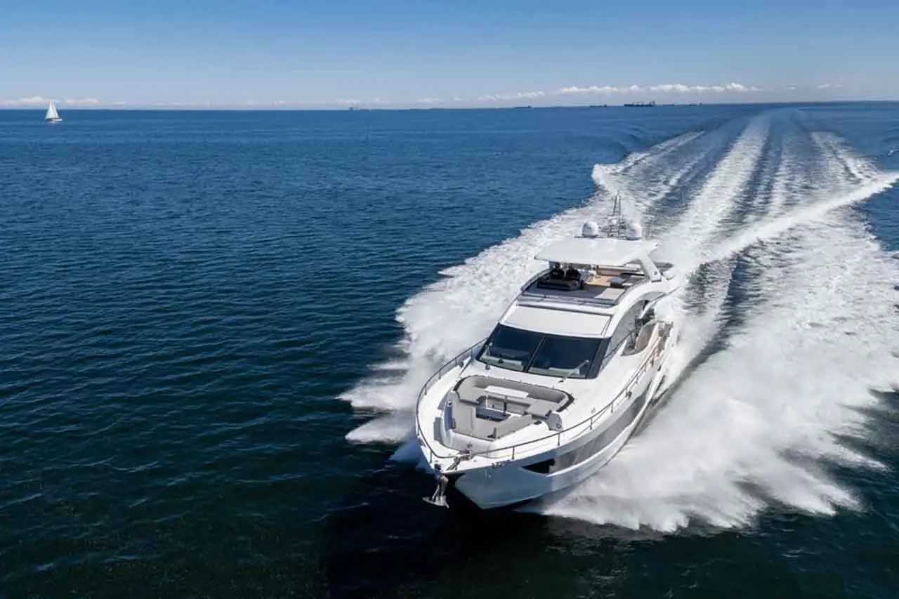 A luxury yacht speeds across the blue sea, creating a white wake, with a sailboat visible in the distance under a clear sky.