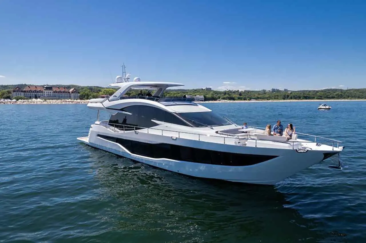 Luxury motor yacht anchored on clear waters with guests relaxing on deck, a beachside hotel visible in the background.