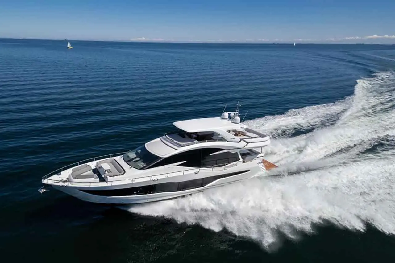 A sleek white yacht cruises through calm waters, leaving a wake against a clear blue sky and distant sailboats.