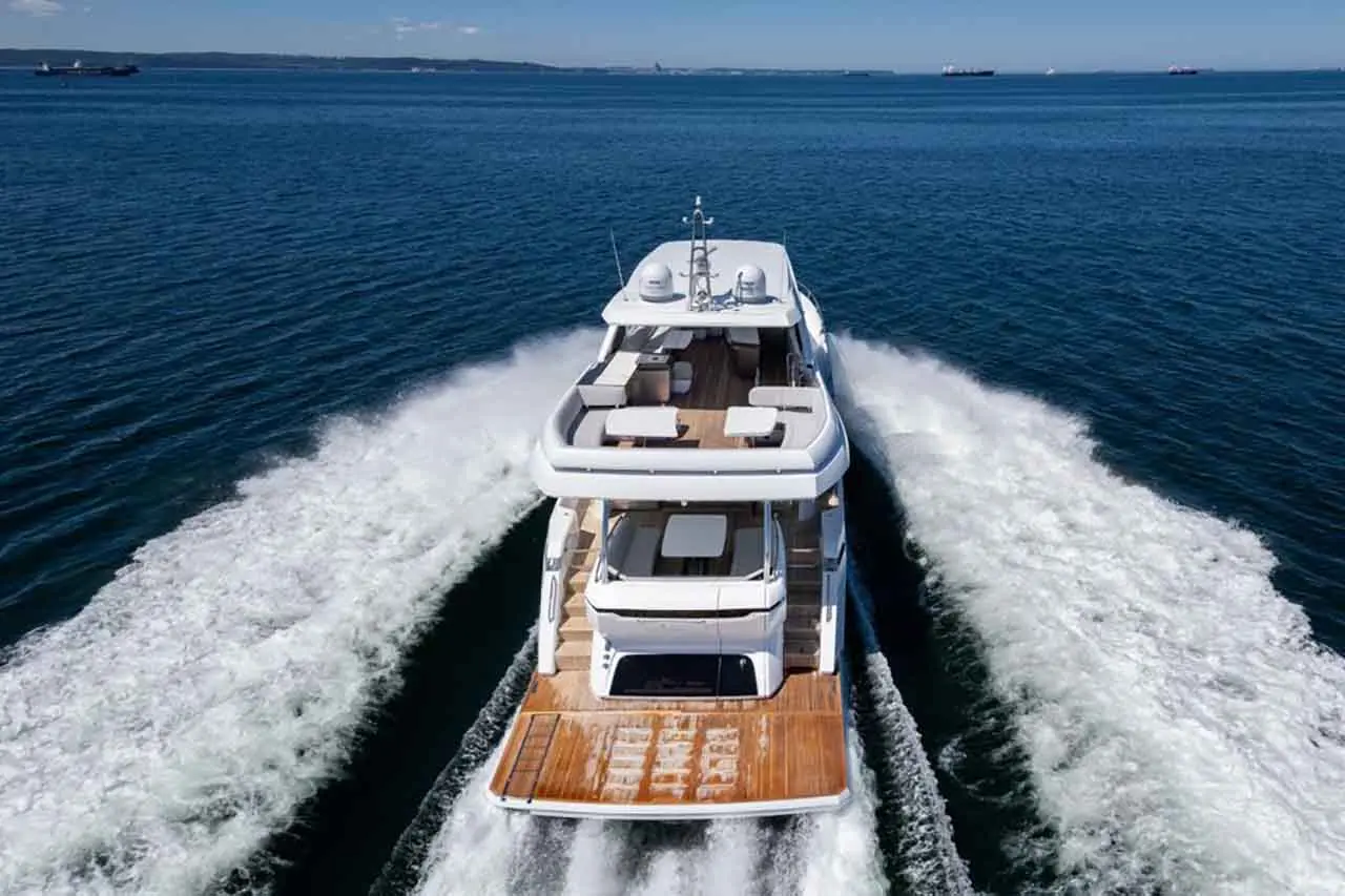 Luxury motor yacht cruising on blue water, leaving a wake, with outdoor seating and a clear sky in the background.