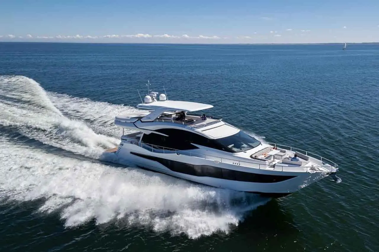 Luxury yacht cruising on clear blue waters, leaving a wake, with a sunny sky and distant sailboats in view.