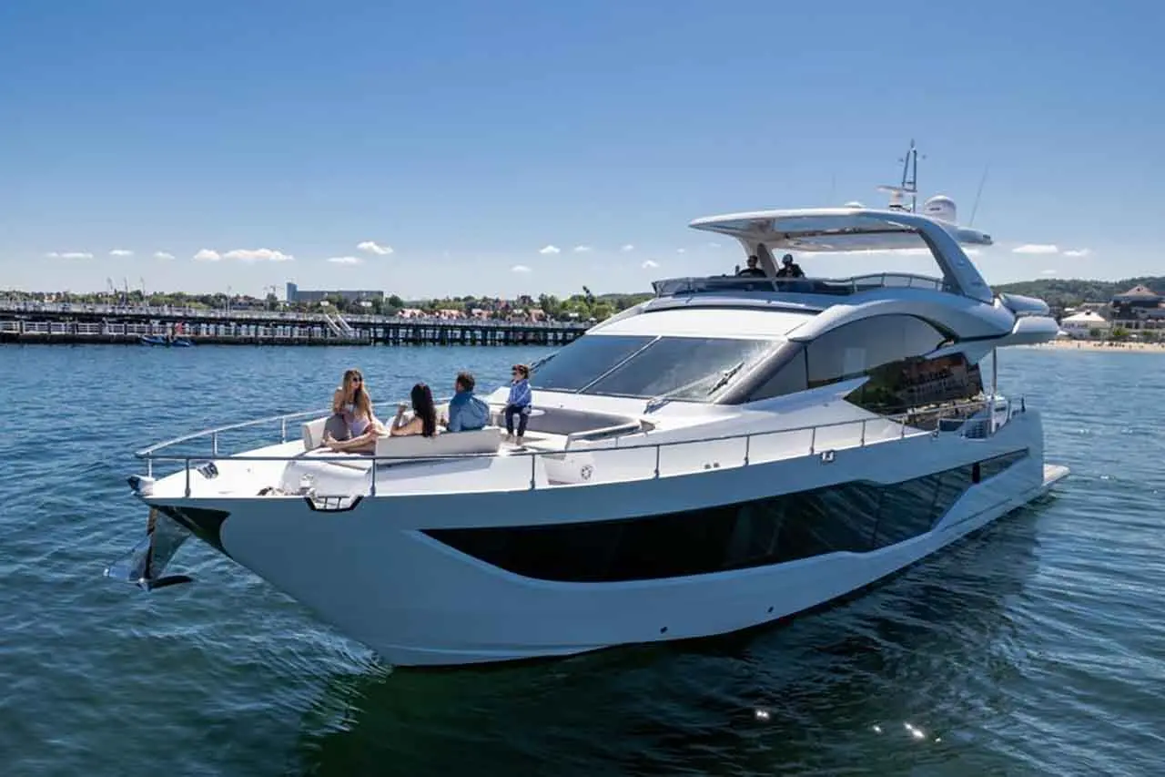 A luxury yacht with guests enjoying the sun on the deck, moored in calm waters with a dock and buildings in the background.