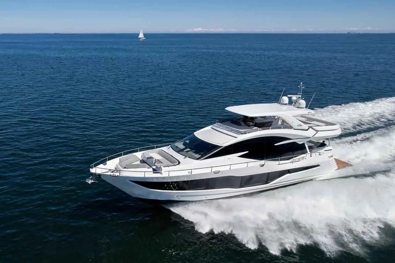 A sleek white yacht glides through calm blue waters, creating frothy waves, with a distant sailboat visible on the horizon.