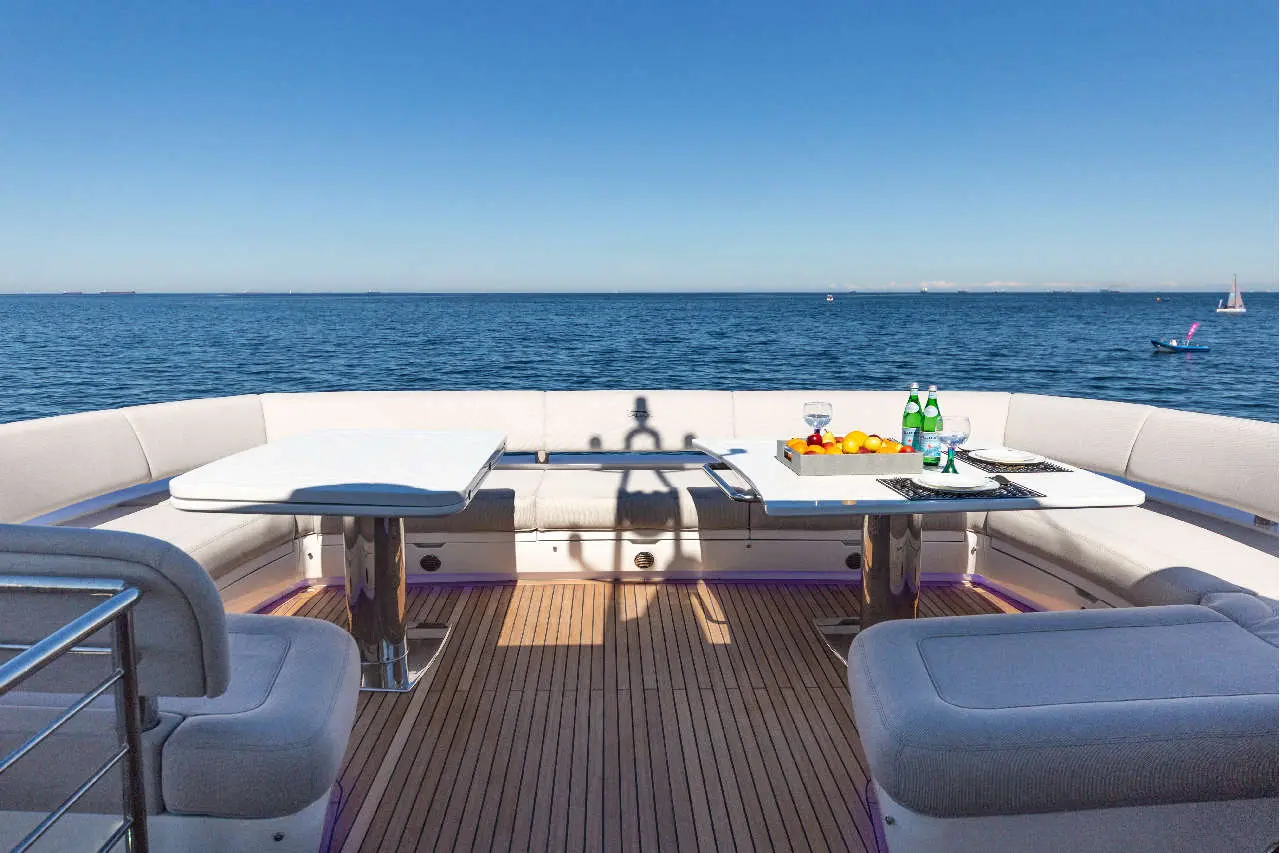 Luxury yacht deck with two tables, fruit platter, drinks, and scenic ocean view under clear blue sky.