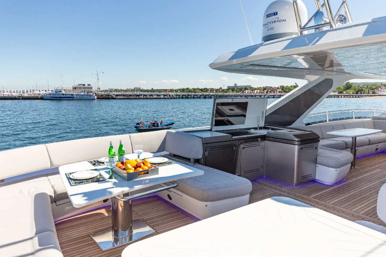 Luxurious yacht deck with a table set for dining, featuring drinks and fruit, overlooking a calm harbour with boats.