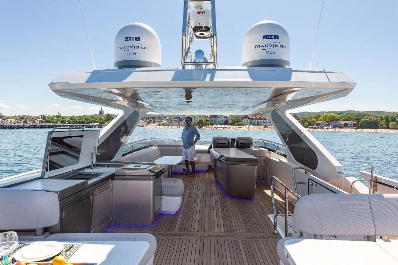 A man stands on the deck of a luxury yacht, featuring modern furniture and ocean views, with a TracVision satellite dish above.